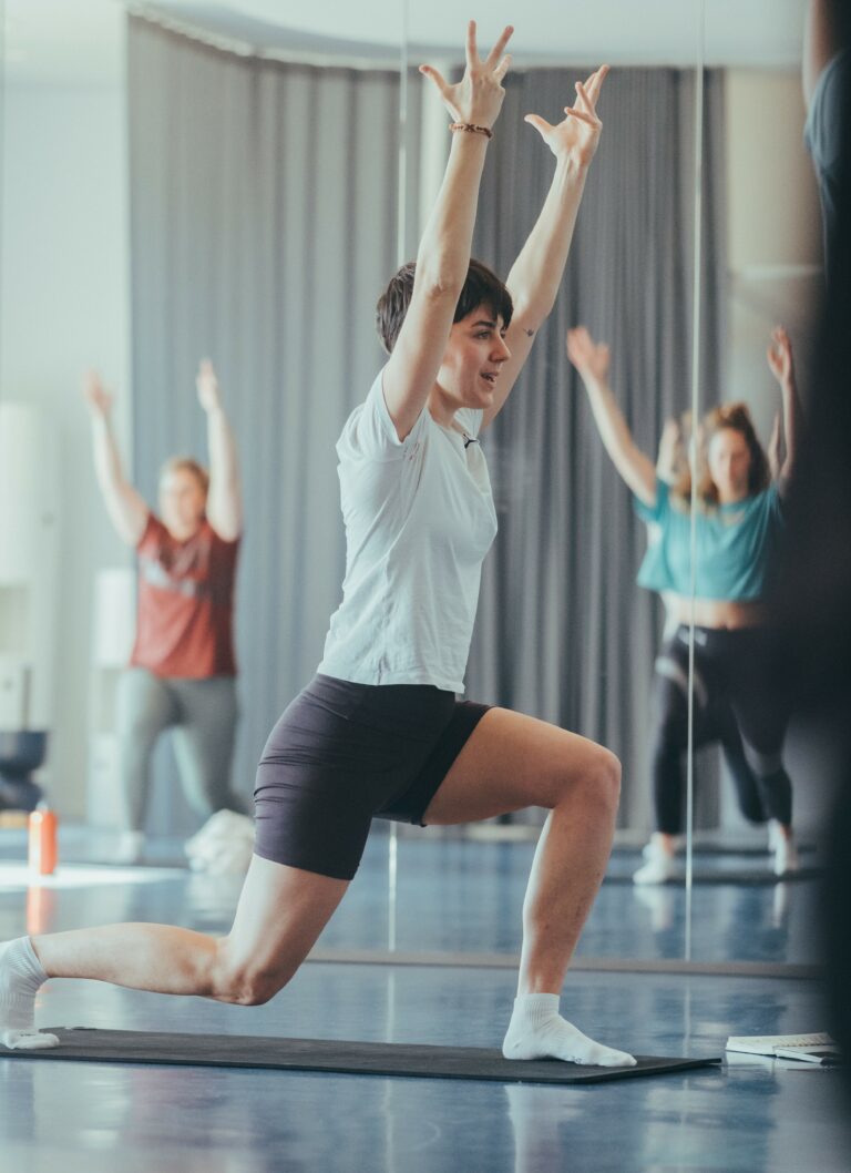Une femme aux cheveux courts, professeure de Pilates, en position de fente haute avec les bras levés, dans un studio avec un miroir en arrière-plan.